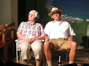 Emilia (M. Hornbogner) und Korbinian (H. Mummert) sitzen im Garten, während Nachbar Schmittke ein Sommerfest feiert. © OVTP / mib